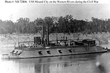 Black and white photo shows a warship with a sloped casemate and two smokestacks. There are four broadside guns and three guns forward.