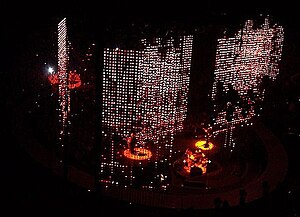 A darkened stage seen from above. Curtains of red and white lights hang down behind and to the sides of the four band members who are lit up with spotlights. Three of the members are on the main stage while the fourth is on the b-stage, an elliptical catwalk that surrounds the main stage.