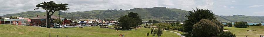 A panoramic view of the town, showing the main shopping strip on the Great Ocean Road (Collingwood Street), the foreshore reserve with surrounding hills in background, and the Apollo Bay Golf Club backed by the beach and bay