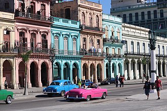 Улица со зданиями, окрашенными в синий, розовый или белый цвет, с автомобилями на переднем плане.