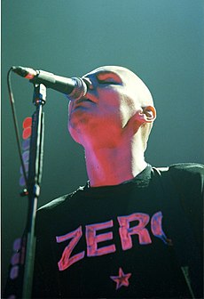 Billy Corgan is depicted from a worm's eye view as he sings into a microphone. He is wearing one of his black "ZERO" T-shirts with lettering and a five-pointed star in reflective silver.