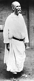 Photographie en noir et blanc représentant un vieil homme debout, dans une tenue d'ermite.
