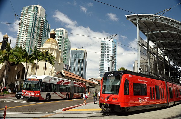 A Rapid bus departs Santa Fe Depot station (left) while a Blue Line train of the San Diego Trolley loads passengers at America Plaza station. The stations are a major MTS hub in Downtown San Diego.