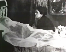 Photographie en noir et blanc d'une femme morte allongée sur un lit, de profil, couverte d'un voile blanc.