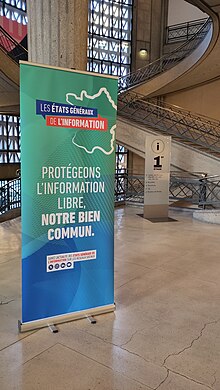 Sur fond de salle des colonnes en béton, un kakémono turquoise annonce les États généraux de l'information et le texte « Protégeons l'information libre, notre bien commun ».