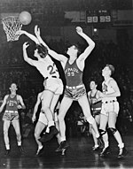 Due giocatori di basket stanno inseguendo una palla da basket, mentre altri quattro giocatori stanno guardando da dietro.