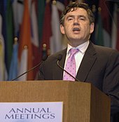 Gordon Brown standing at a podium. Text on the podium states "ANNUAL MEETINGS". A number of flags hang in the background.