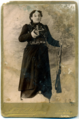 A 1910 postcard (front) portraying the widow of Kevork Chavush, Heghine. On the front of the postcard is the black-and-white image of a woman in a long black dress; she’s holding a handgun in her right hand and a rifle in her left. Bandoliers are wrapped across her chest and around her waist. Nearly lost among the bullets and leather is a round brooch or medallion above her left breast.