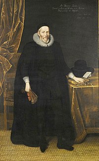 A tall elderly man with a beard, wearing long black robes and a large white ruff. He is standing with a fan in his right hand and with his left hand resting on books on a table.