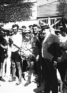 Photographie en noir et blanc d'un cycliste tenant un bouquet, entouré par la foule.