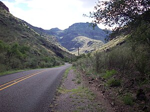 RM 1832, a two-lane road, winds through a canyon with steep hillsides on both sides of the road. In the distance are rugged mountains with steep cliffs covered with desert grasses and brush.