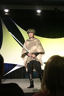 Homme portant un casque, une cape de camouflage et une réplique de sabre laser