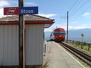 Wooden structure on concrete platform; red train approaches