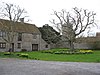 Tickenham Court in England: a large, two-storey stone building with gabled roof and surrounding stone wall. The house has a gravel driveway and large grassy space in front with yellow daffodils and two bare trees.