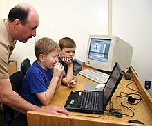 Children being taught how to use a laptop computer in 2005. An older (1990s-era) desktop personal computer's CRT monitor is visible in the background. US Navy 050210-N-2802K-001 Chief Aviation Warfare Systems Operator Richard McCurdy demonstrates how to create a PowerPoint presentation to Shirley Lanham Elementary School fourth-graders.jpg