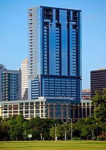 A view from Zilker Park of the building; the tower is topped out; the building is rectangular, and there are no setbacks; the building has a reflective blue facade; the tower appears to look like a tall rectangle stacked on top of another smaller rectangle.