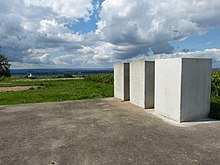 Tři betonové bloky stojí na betonové podložce, obklopené zvlněnou travnatou půdou a kukuřičnými poli. V dálce je vidět kostelní věž.