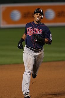 Un hombre con una camiseta de béisbol azul, pantalones grises a rayas, un casco de bateo y guantes negros corre por las bases.