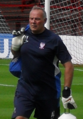 A man with grey hair is wearing a dar blue top. He is standing on a grass field.