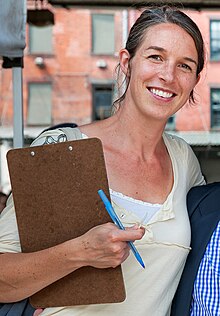 Anne Saxelby holding a pen and a clipboard, smiling and facing the camera.