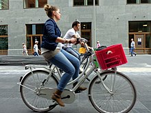 The angled-back handlebars (woman in the foreground) allow an erect sitting position increasing comfort, in contrast with straight handlebars (man in the background) which increase tensions on the lumbar area and shoulders. Red eurobox as bicycle basket. Bicycle in The Hague 39.JPG