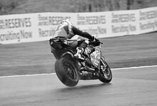 Black and white image of a racing motorcycle taken from a three quarter rear camera position, with the motorcycle rear wheel elevating when braking hard for a left bend on a wet track