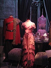 Le costume au centre de la photo est une robe rose à volants, et l'autre, sur la gauche, est une tunique rouge avec une ceinture et un manteau en fourrure placé sur une épaule.