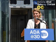 Feinberg hinter einem Rednerpult mit der Aufschrift „ADA@30“ zu 30 Jahren Gesetzgebung zur Barrierefreiheit, hinter ihr ein neuer Aufzug am so beschilderten U-Bahnhof „Astoria Boulevard“
