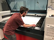A technologist inserts a sheet of acrylic into a commercial grade laser cutter as part of the manufacturing process to create face shields