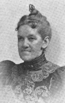 A white woman, hair dressed to the crown, wearing a high-collared dark dress with elaborate lace trim on the bodice, and puffed sleeves