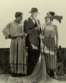 A man in a suit stands between a man and women dressed in costume as figures from the Roman Empire