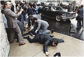 Photographie en couleur. Sur un trottoir, deux hommes sont au sol. Un homme tient un uzi dans les mains prêt à tirer.