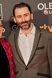 A bearded man wearing a brown suit with a white shirt.