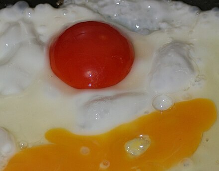 A chicken egg frying with an extremely thick red yolk. A normal-coloured yolk can also be seen, having been accidentally burst during the frying process.