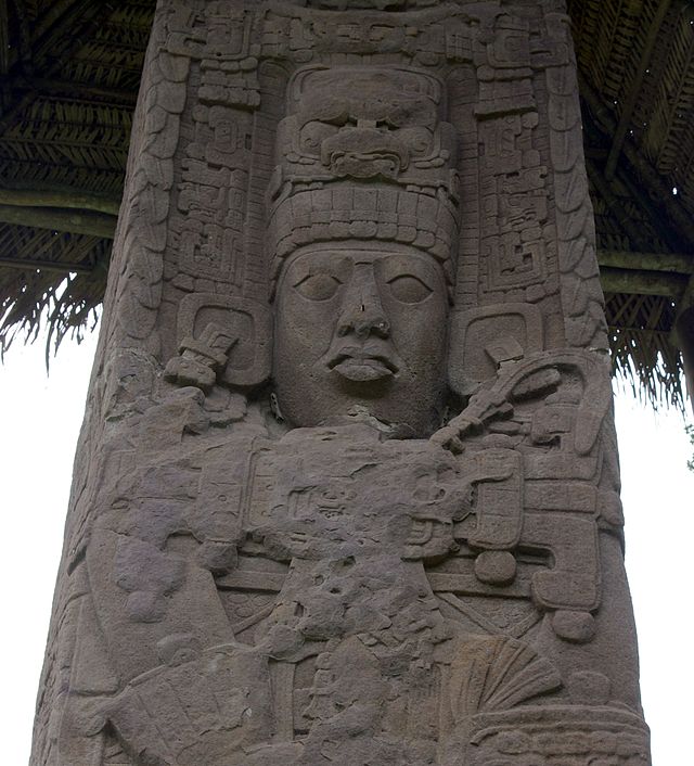 Close up of upper portion of an intricately carved stela, showing the face of a king with elaborate headdress and jewellery