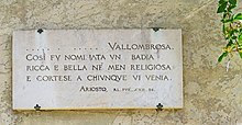 Fotografia barwna, jasna marmurowa płyta z cytatem po włosku przymocowana do ściany, z prawej strony zwisa kawałek rośliny