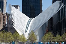 A curved white structure with projecting ribs at an angle, seen with budding trees in front and modern high-rises behind it in sunlight with a clear blue sky behind