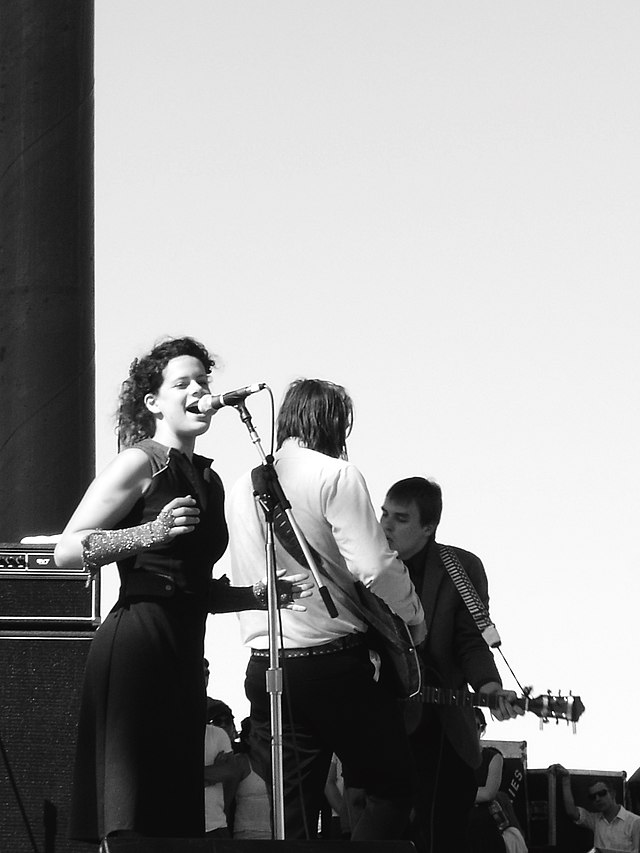 Plan large, photo en noir et blanc : Régine Chassagne en robe noire ajustée chantant sur scène.