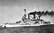 A large, light gray battleship plows through the water at high speed. Thick black smoke pours from the two round smoke stacks