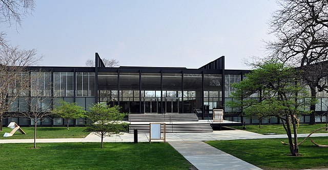 a low glass and steel building behind a sidewalk and small lawn and three trees