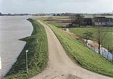 A river dike with a narrow road on top, high water levels on the river to the left, low lying meadows and a farm on the right