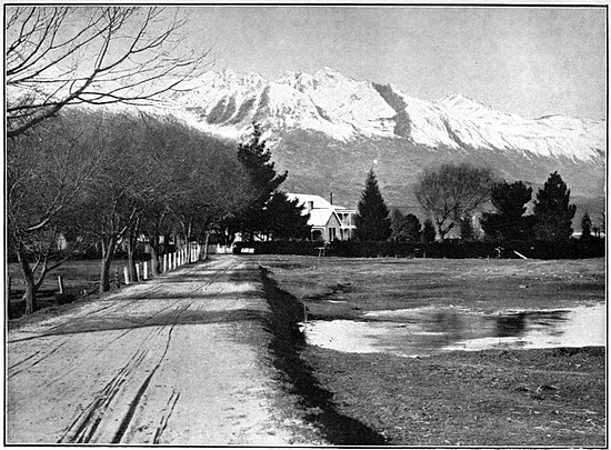 A rutted road beside a lake with a large house obscured by pine trees in the distance and a tall snowy mountain range behind.