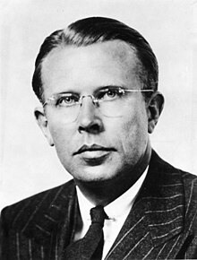 Head and shoulders of a man wearing rimless glasses, and a dark suit and tie