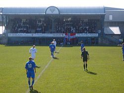 Pertandingan sepak bola dengan pemain dan wasit di lapangan. Kecil penonton berdiri penuh dengan orang-orang di latar belakang.