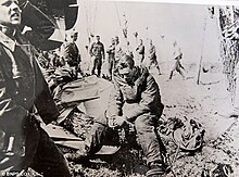 Photo noir et blanc d'un homme hagard assis sur les restes d'un avion. Autour de lui, beaucoup d'hommes en uniformes marchent ou travaillent.