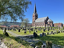 Foto einer mit roten Backsteinen erbauten Kircher, im Vordergrund ein Friedhof