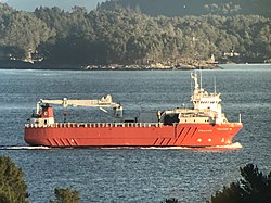 Sideprofil av rødt lasteskip som seiler inne i en fjord. Fint vær med fastland på ene siden og holmer på andre siden av skipet.
