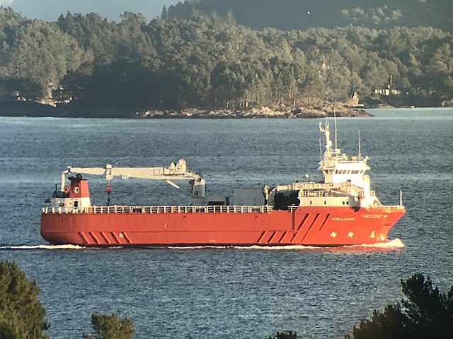 Sideprofil av rødt lasteskip som seiler inne i en fjord. Fint vær med fastland på ene siden og holmer på andre siden av skipet.