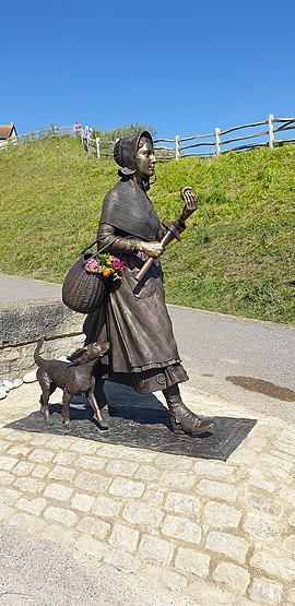 A bronze statue of Mary Anning. She is walking, holding an ammonite with a basket over one arm and her dog trailing behind.