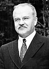 A man in a dark suit, light shirt and dark tie, smiling